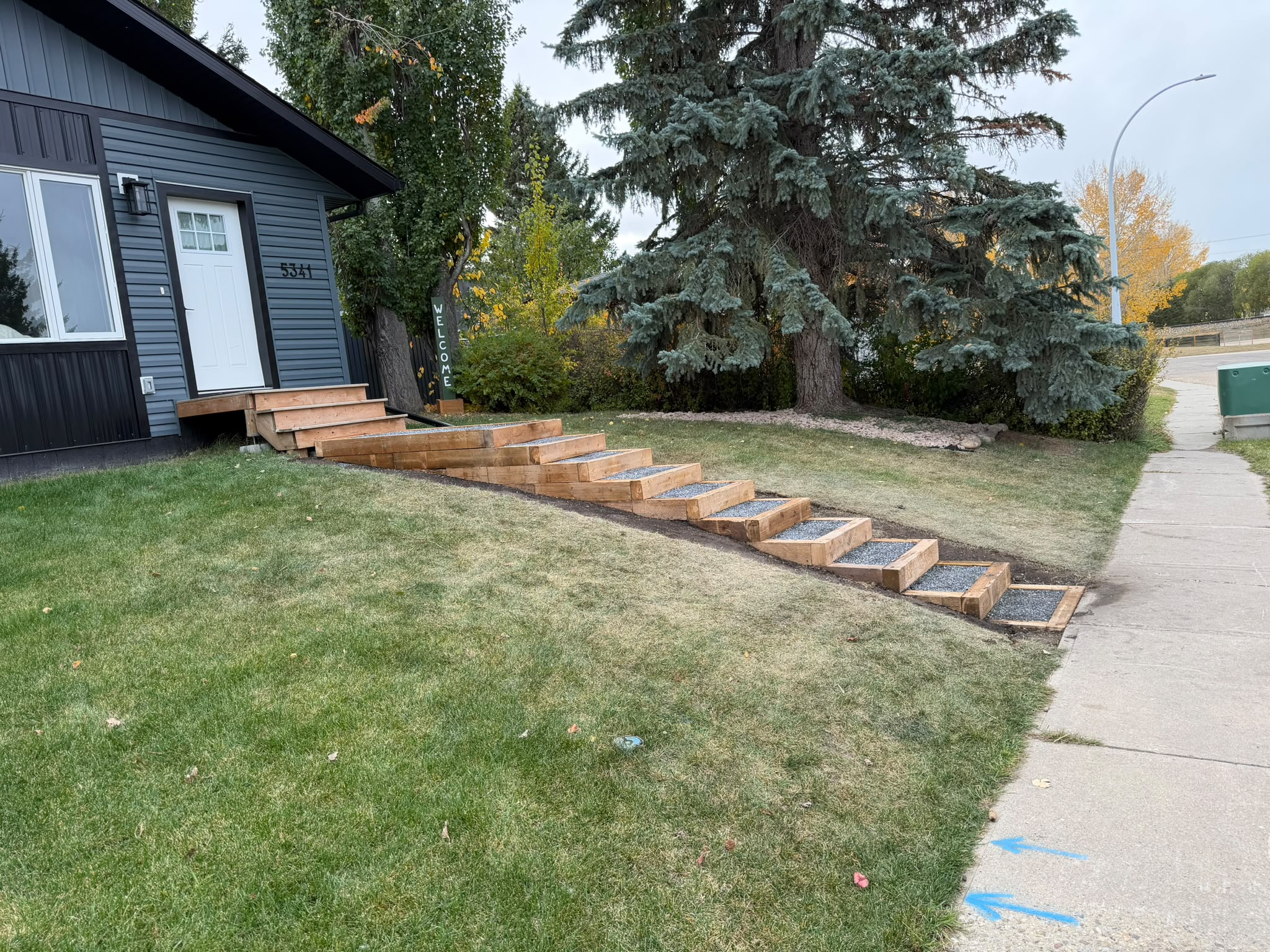 Landscape tie steps with crushed bluestone treads climbing a front lawn hill from the sidewalk up to the existing front steps of a home in Innisfail, AB, built by Green Ladder Contractors